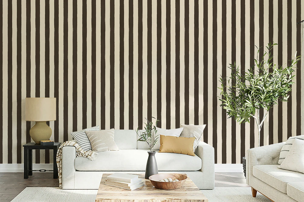 A styled living room featuring a prominent wall with vertical dark brown and beige stripes, decorated with a white sofa, a wooden coffee table, a side table with a lamp, and a potted green tree.