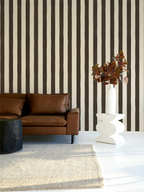 A modern living room scene featuring a brown leather sofa against a wall with bold vertical brown and cream stripes. A white sculptural side table holding a tall white vase with dried brown leaves sits on a white floor next to a beige woven rug.