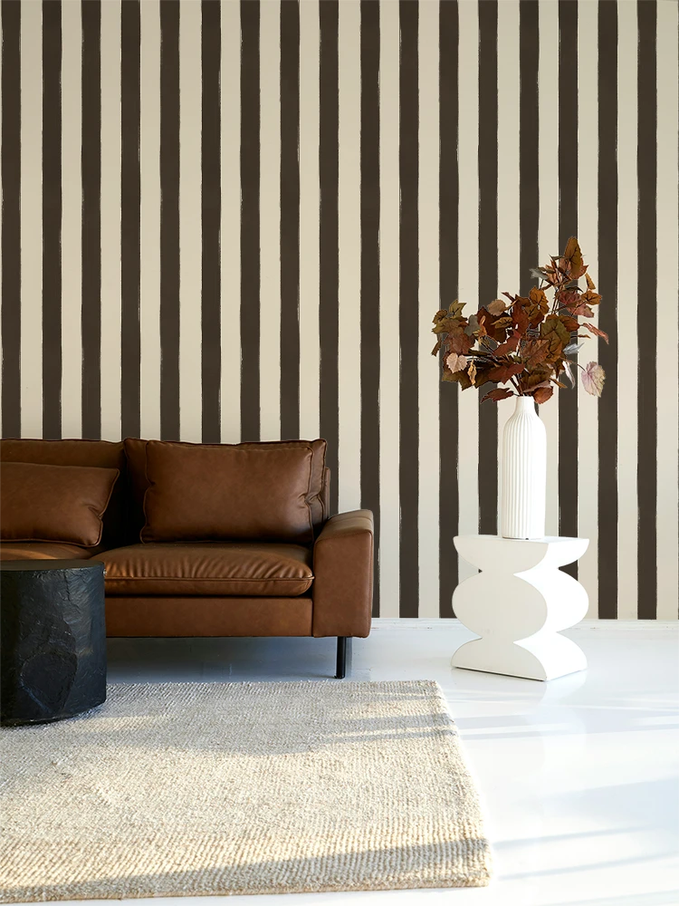 A modern living room scene featuring a brown leather sofa against a wall with bold vertical brown and cream stripes. A white sculptural side table holding a tall white vase with dried brown leaves sits on a white floor next to a beige woven rug.