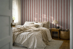 Bedroom with white and brown sheets, a wooden floor, nightstand with a lamp, and a striped wallpaper with pink brush strokes. 