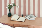 A small desk with a vase with white roses, a notepad and a pen, three books, and a striped wallpaper with pink brush strokes. 