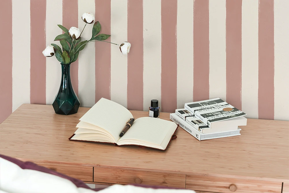 A small desk with a vase with white roses, a notepad and a pen, three books, and a striped wallpaper with pink brush strokes. 
