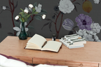 Open book on a wooden desk with stationery items, with chinoiserie wallpaper on the wall. 