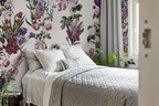 A styled bedroom featuring a bed with white bedding and a gray quilted coverlet, set against a wall with white wallpaper decorated with large pink, purple, and green floral prints. Gray curtains hang by a window, and a green plant sits on the floor.
