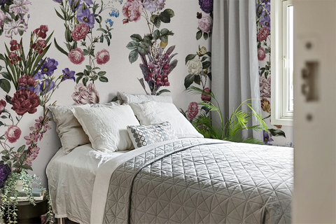 A styled bedroom featuring a bed with white bedding and a gray quilted coverlet, set against a wall with white wallpaper decorated with large pink, purple, and green floral prints. Gray curtains hang by a window, and a green plant sits on the floor.