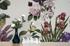 A styled interior scene showing a large botanical wallpeper mural with illustrated flowers and leaves in soft pink, purple, green, and burgundy tones. In front of the wall is a light wood surface holding a dark blue vase with white flowers and a stack of books.