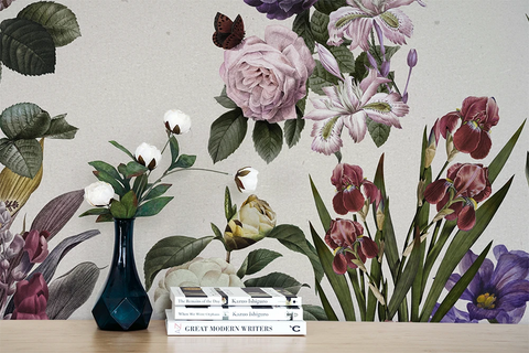 A styled interior scene showing a large botanical wallpeper mural with illustrated flowers and leaves in soft pink, purple, green, and burgundy tones. In front of the wall is a light wood surface holding a dark blue vase with white flowers and a stack of books.