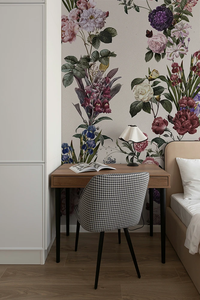 A styled bedroom corner featuring a wooden desk with a black-and-white checkered upholstered chair, a white table lamp, and an open magazine. The background is dominated by a large-scale floral wallpaper with pink, purple, and white flowers and green leaves, set against a light wood floor.