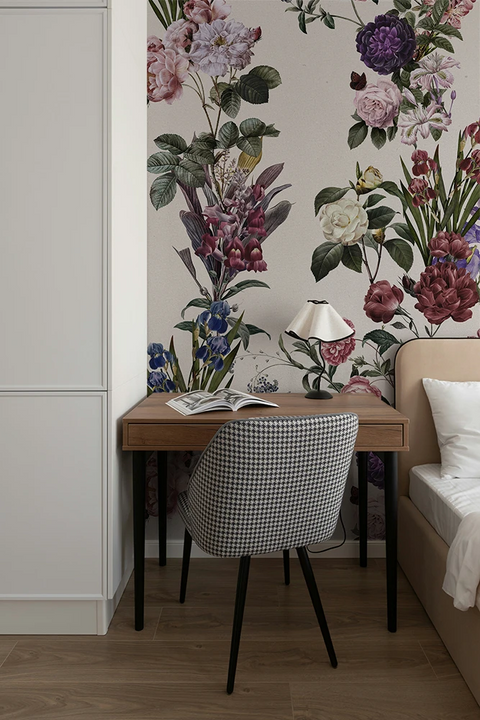 A styled bedroom corner featuring a wooden desk with a black-and-white checkered upholstered chair, a white table lamp, and an open magazine. The background is dominated by a large-scale floral wallpaper with pink, purple, and white flowers and green leaves, set against a light wood floor.
