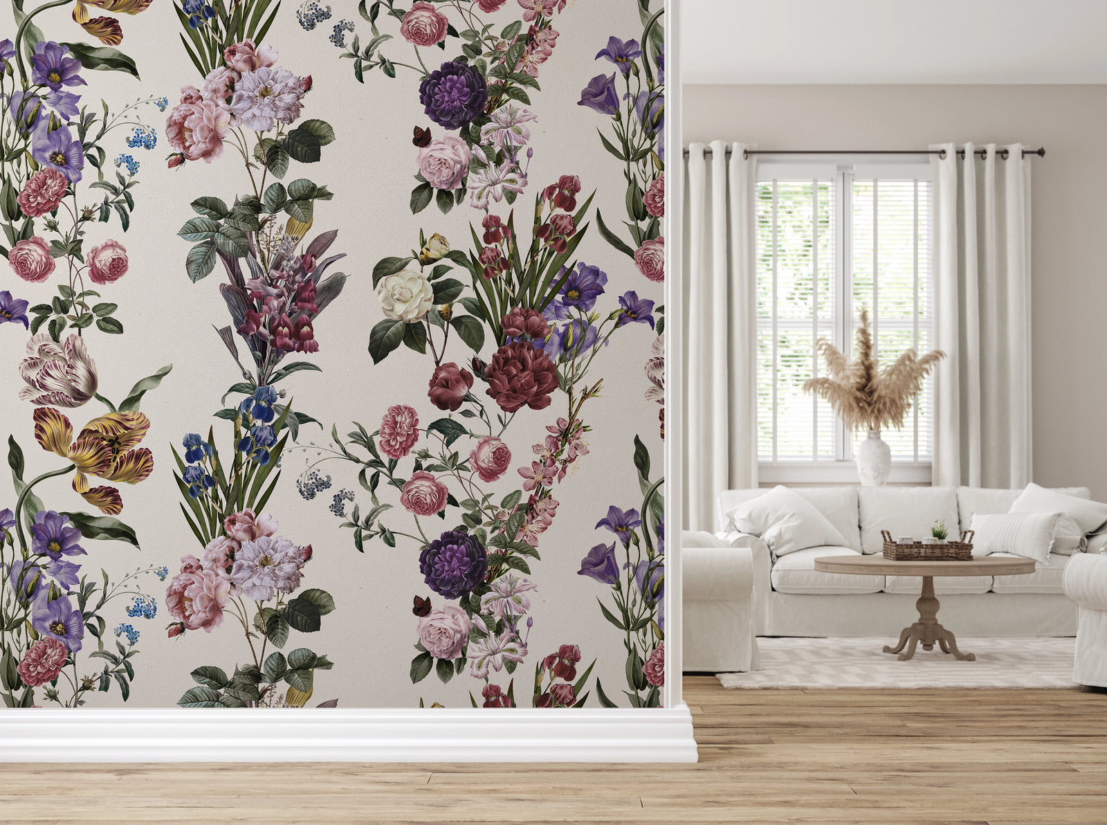 A room interior showing floral wallpaper on the left wall with a white sofa, window, curtains, and wood floor on the right. The wallpaper features a dense botanical pattern with pink, purple, blue, yellow, and white flowers on a warm off-white background.