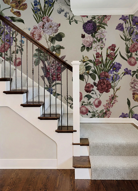 A staircase interior with dark wood steps and a white handrail, set against a wall covered in large floral wallpaper. The wallpaper features a dense botanical pattern with pink, purple, blue, and green flowers and leaves on a light background.