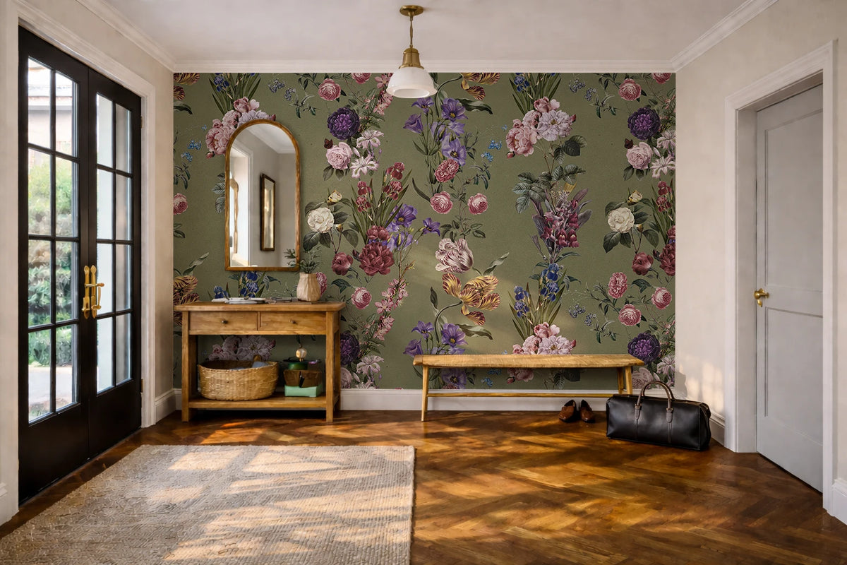 A styled interior room featuring a prominent olive green floral wallpaper with pink, purple, and white botanical patterns on the back wall. The room includes a wooden console table with a mirror, a wooden bench, a black leather bag on the floor, a patterned rug, and a light wood herringbone floor.