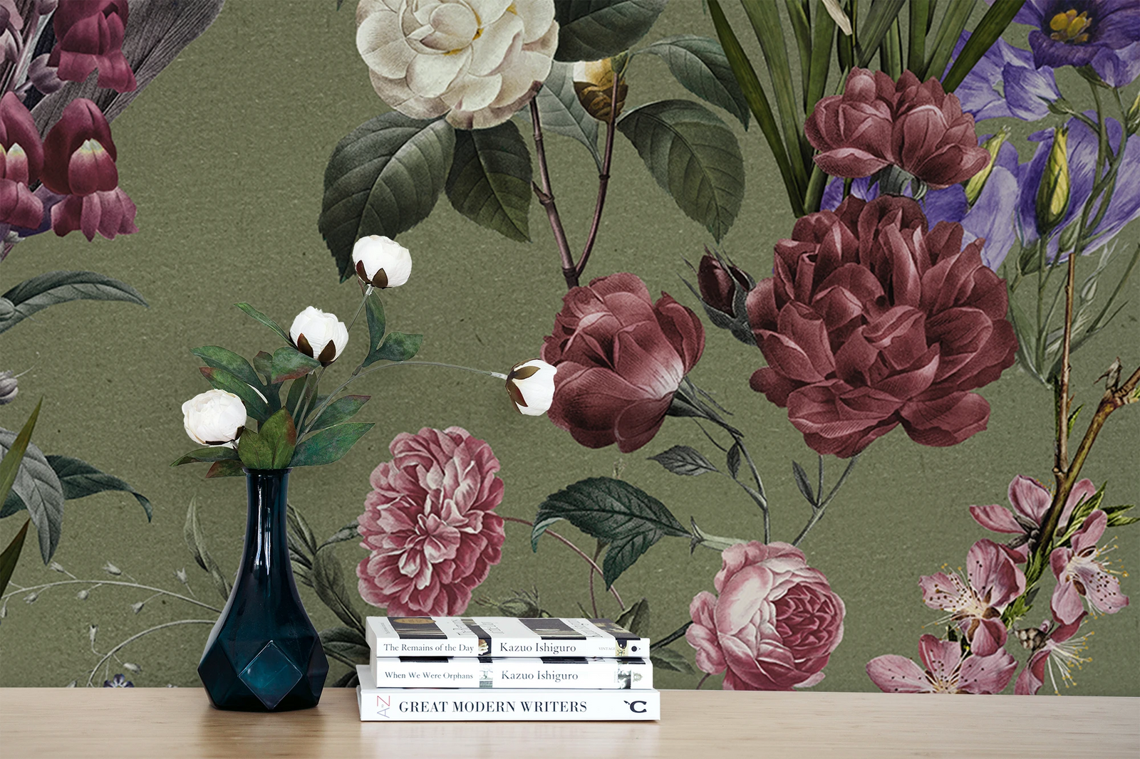 A styled interior scene showing a floral wallpaper with large vintage flowers in burgundy, pink, white, and purple tones on an olive green background. In front of the wall is a light wood surface with a dark blue vase holding white flowers and a stack of books.