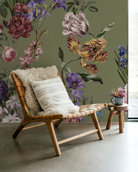 A styled interior scene with a woven wooden lounge chair, cream pillows, a small stool, and a large floral wall mural in muted greens, mauves, purples, and tan tones.