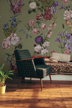 A styled interior scene with a large floral wallpaper mural on an olive green wall, featuring pink, purple, and white flowers with green leaves. In front of the wall is a dark woven armchair, a patterned sofa with pillows, a potted plant on a wooden floor, and a white wall at the side.