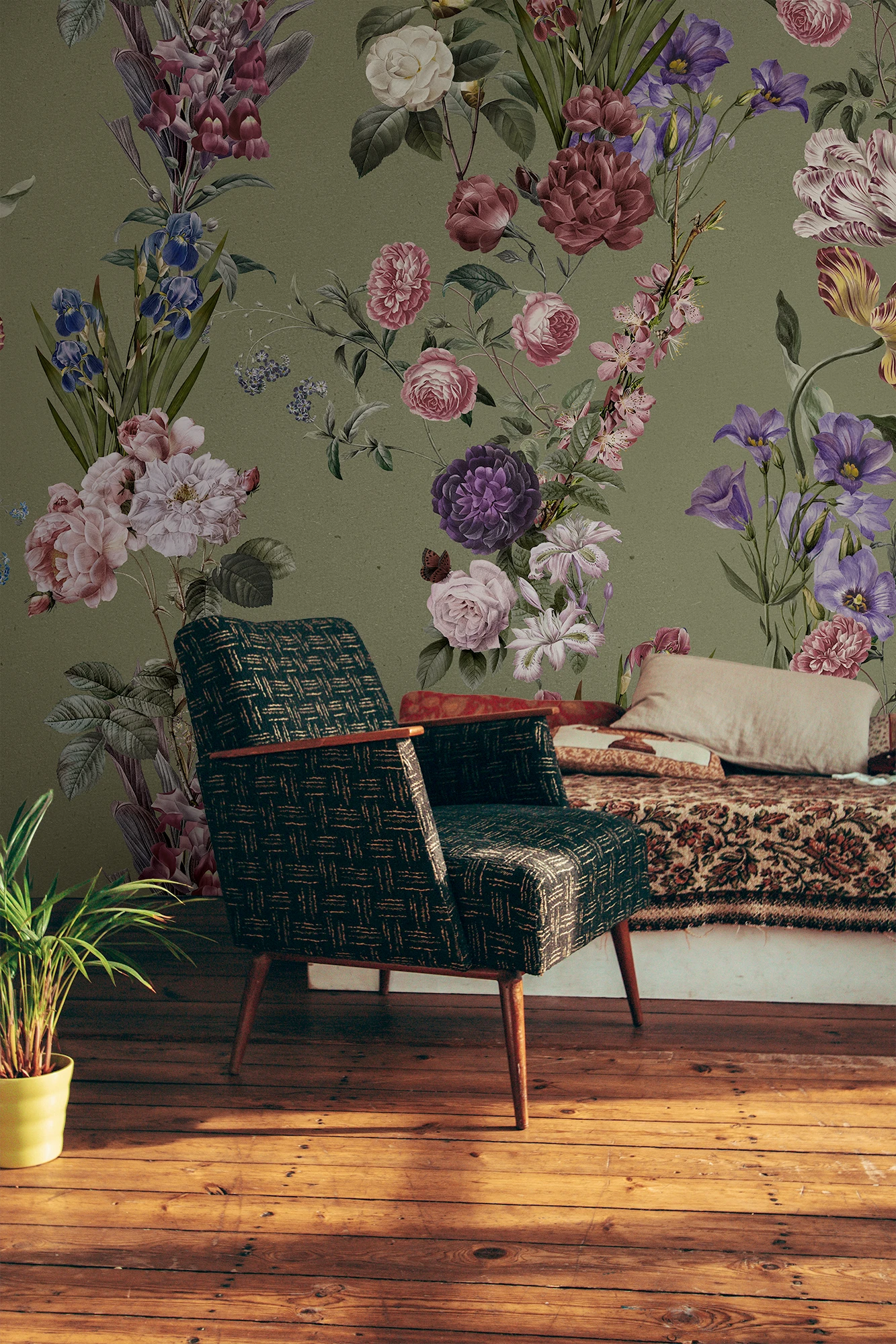 A styled interior scene with a large floral wallpaper mural on an olive green wall, featuring pink, purple, and white flowers with green leaves. In front of the wall is a dark woven armchair, a patterned sofa with pillows, a potted plant on a wooden floor, and a white wall at the side.