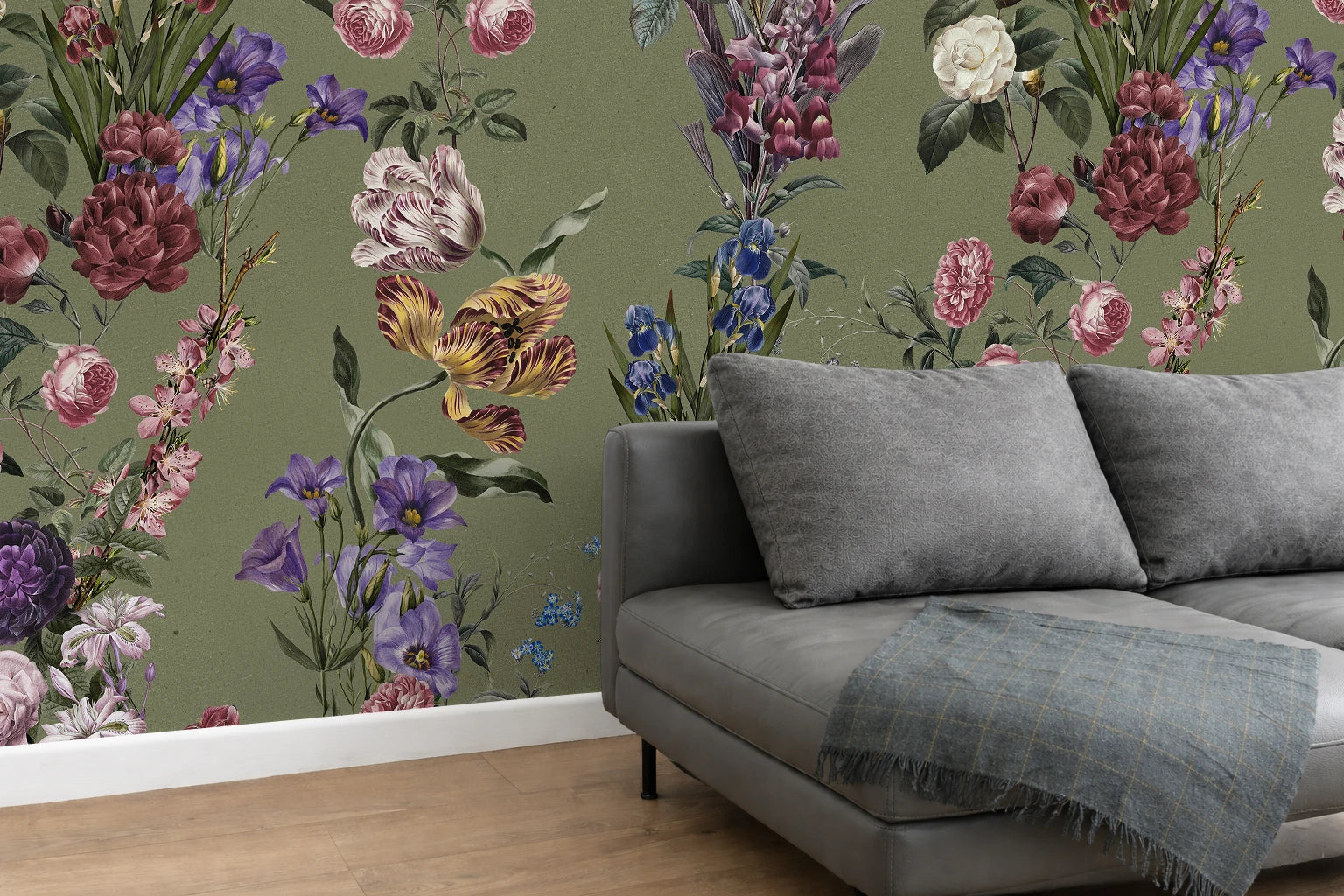 A styled interior scene showing a gray sofa with two gray cushions and a blue-gray throw blanket, set against a wall covered in a dense floral wallpaper with pink, purple, blue, and orange flowers on an olive green background.