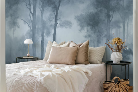 A styled bedroom featuring a large wallpaper mural with a misty forest scene in muted dusty-blue tones. The bed is dressed in cream and beige bedding with multiple pillows, flanked by two black nightstands holding a white table lamp and a white vase with dried flowers, and a woven basket bag sits on the floor.