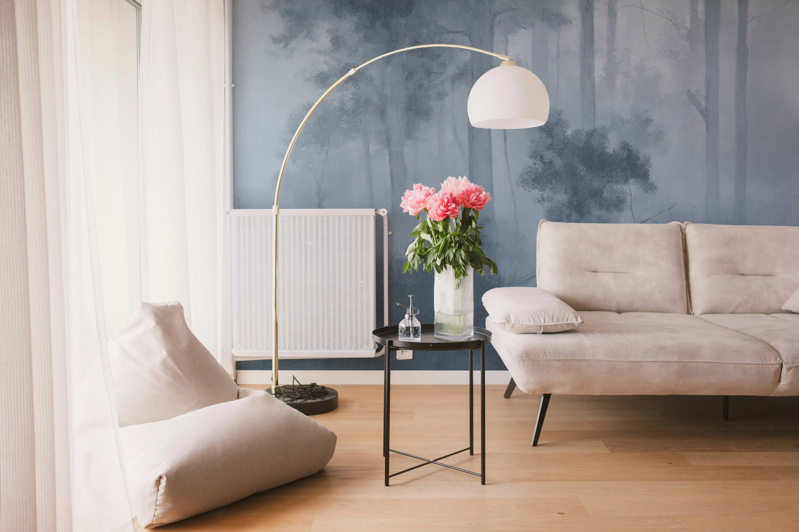 A modern living room scene featuring a beige fabric sofa, a matching floor cushion, a black side table with a vase of pink flowers, a gold arched floor lamp, and a blue forest-patterned accent wall.