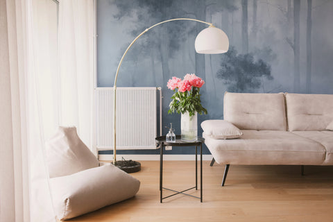 A modern living room scene featuring a beige fabric sofa, a matching floor cushion, a black side table with a vase of pink flowers, a gold arched floor lamp, and a blue forest-patterned accent wall.