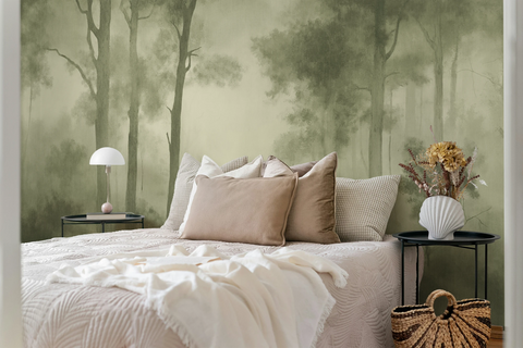 A styled bedroom scene featuring a bed with beige and cream bedding, set against a large olive colored forest mural wallpaper. Two black side tables flank the bed, one holding a white dome lamp and the other a white vase with dried flowers, with a woven straw bag on the floor.