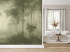 A large wallpaper mural featuring a misty forest scene in muted olive and sage green tones, shown installed on a wall in a bright, modern living room with a white sofa, window, and light wood floor.