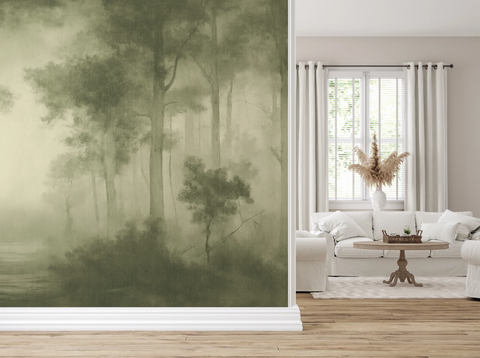 A large wallpaper mural featuring a misty forest scene in muted olive and sage green tones, shown installed on a wall in a bright, modern living room with a white sofa, window, and light wood floor.