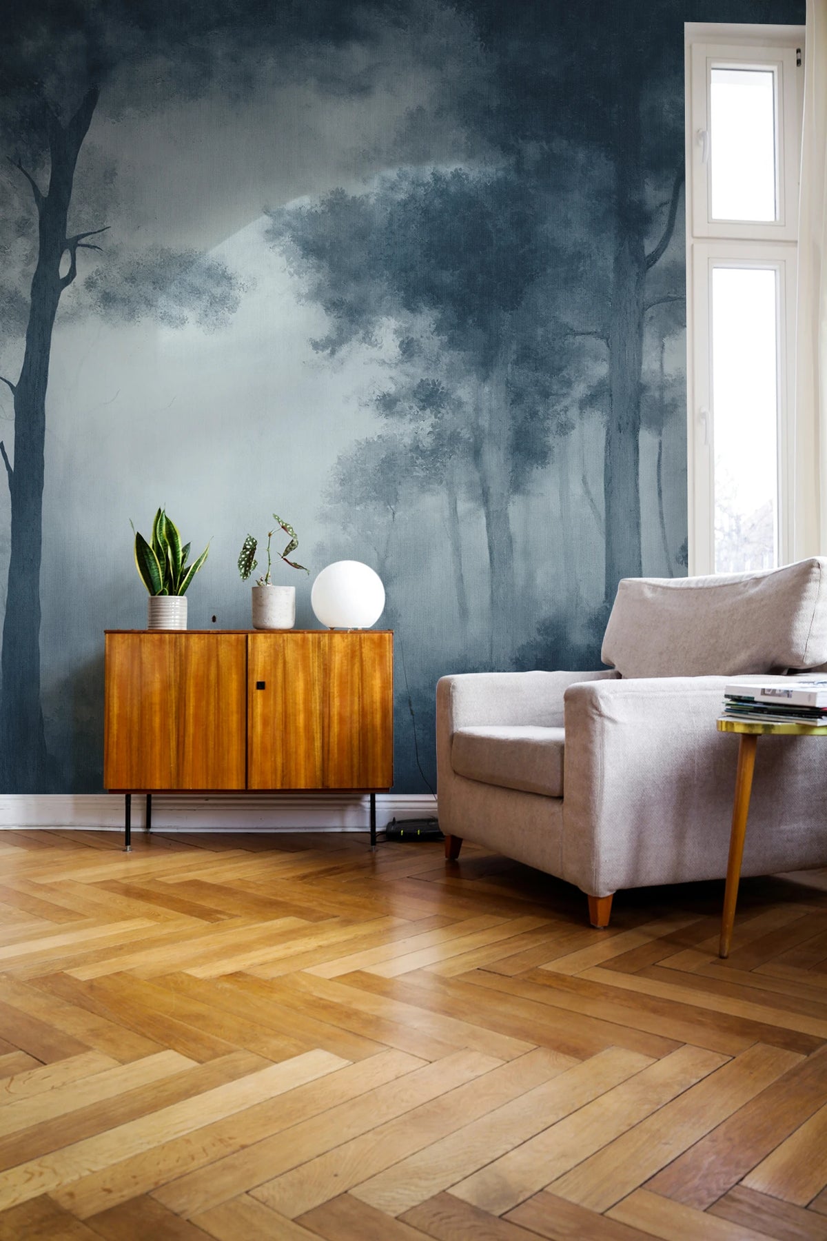 A styled living room corner featuring a dark blue and gray misty forest mural wallpaper on the wall, paired with light wood herringbone flooring, a mid-century wooden cabinet, a beige armchair, and a tall window.