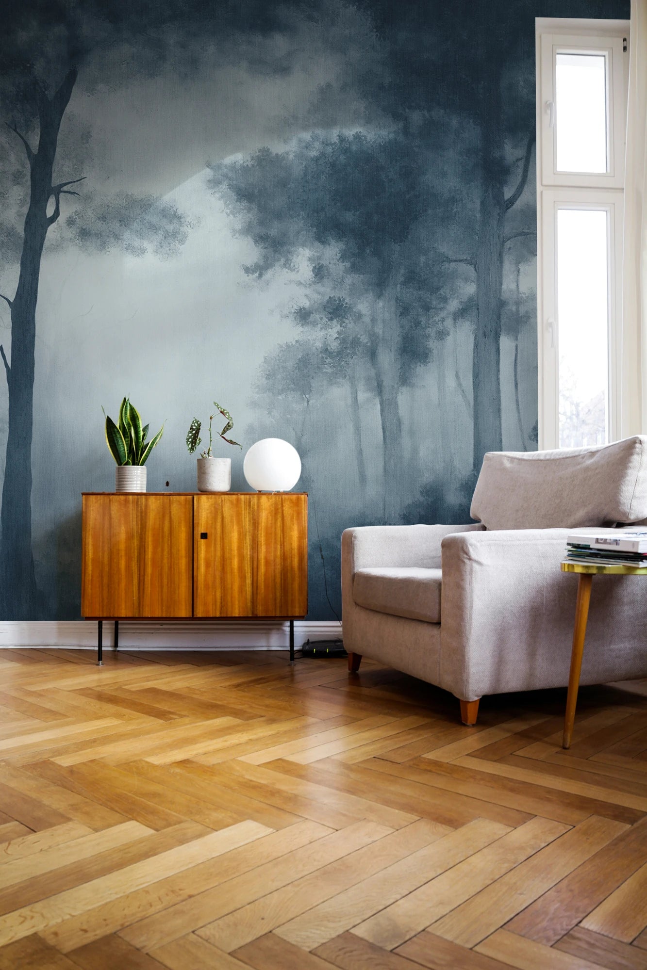 A styled living room corner featuring a dark blue and gray misty forest mural wallpaper on the wall, paired with light wood herringbone flooring, a mid-century wooden cabinet, a beige armchair, and a tall window.