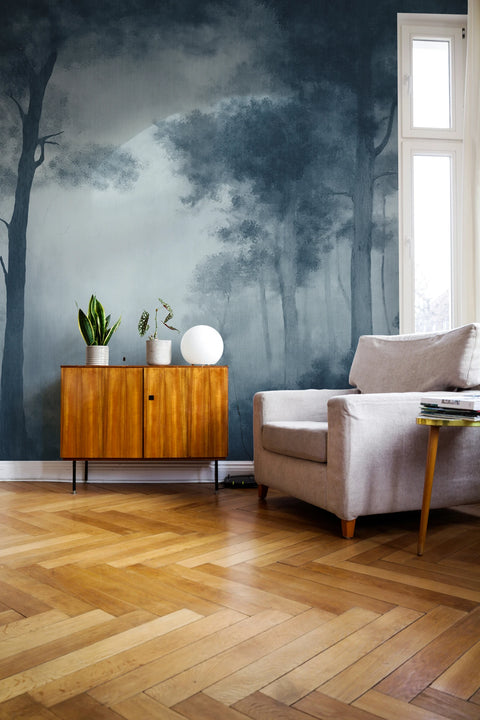 A styled living room corner featuring a dark blue and gray misty forest mural wallpaper on the wall, paired with light wood herringbone flooring, a mid-century wooden cabinet, a beige armchair, and a tall window.
