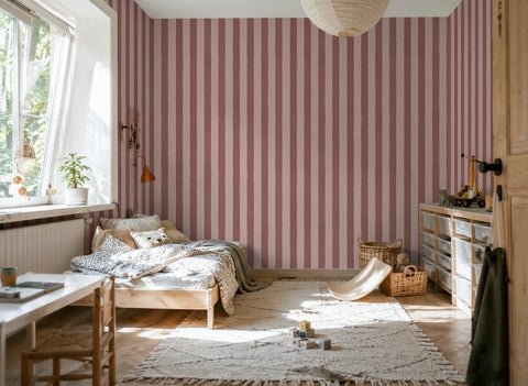 A styled bedroom interior featuring prominent vertical pink striped wallpaper on the main wall, with a wooden bed, light-colored rug, storage cabinet, and window on the left.