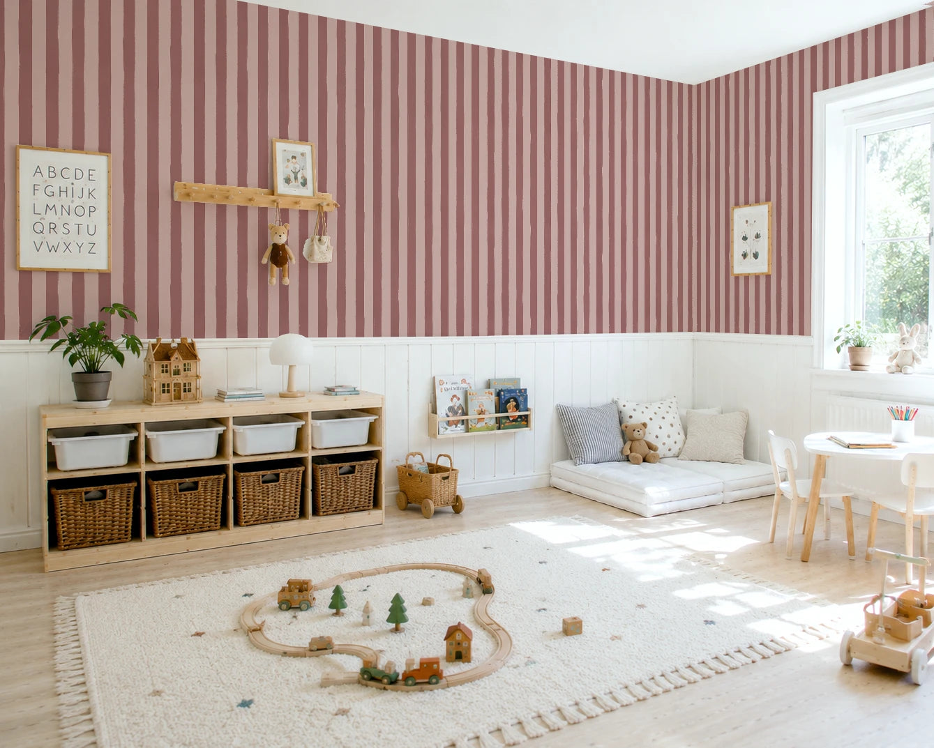 A styled children's playroom featuring dusty rose and white vertical striped wallpaper, a light wood credenza with woven baskets, a white rug with a wooden toy train track, and a small white table with chairs near a bright window.