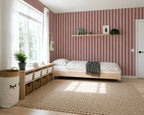 A styled children's bedroom featuring a light wood single bed with a dark green throw blanket, set against a wall of pink and burgundy vertical striped wallpaper. A matching striped area rug covers the light wood floor, and a white storage bench with woven baskets sits next to a large window.