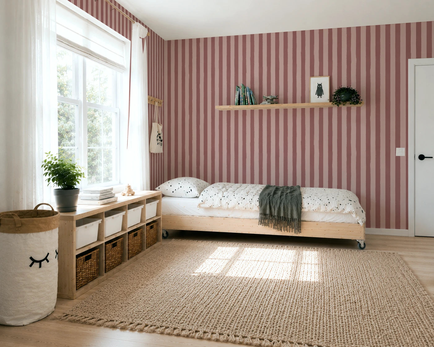 A styled children's bedroom featuring a light wood single bed with a dark green throw blanket, set against a wall of pink and burgundy vertical striped wallpaper. A matching striped area rug covers the light wood floor, and a white storage bench with woven baskets sits next to a large window.