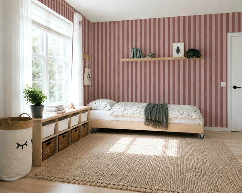 A styled children's bedroom featuring a light wood single bed with a dark green throw blanket, set against a wall of pink and burgundy vertical striped wallpaper. A matching striped area rug covers the light wood floor, and a white storage bench with woven baskets sits next to a large window.
