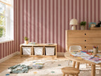 A styled children's room featuring pink and burgundy vertical striped wallpaper, a light wood storage bench with white bins, a wooden dresser, a small round wooden table with art supplies, and a beige rug.