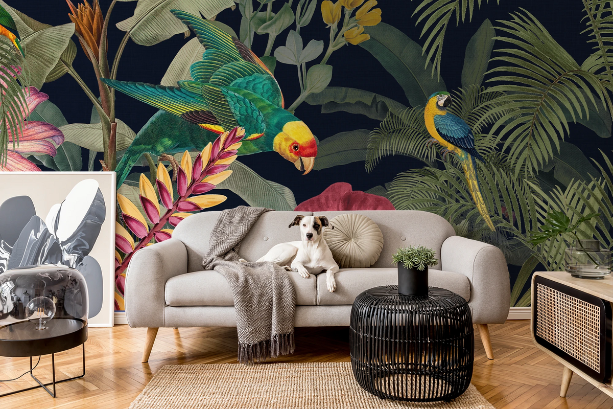 A styled living room featuring a large tropical jungle wallpaper mural with parrots and palm leaves. In front of the wall is a light gray sofa with a dog lying on it, a gray throw blanket, a round beige pillow, a black woven coffee table, and a light wood side table with a plant.