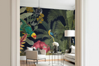 A styled interior room showing a large tropical wallpaper mural with dark navy background, green palm leaves, colorful birds, and pink flowers. The mural is installed behind a light beige armchair, a small round table with flowers, and a white sofa, framed by cream door trim.