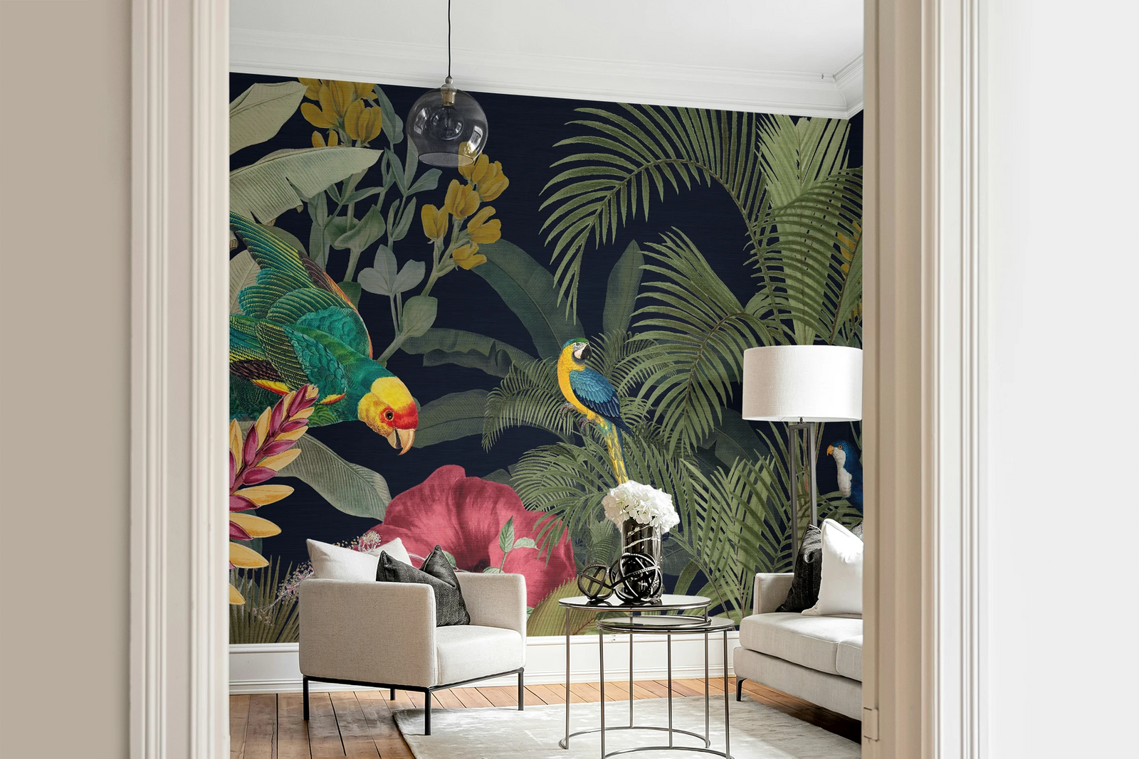 A styled interior room showing a large tropical wallpaper mural with dark navy background, green palm leaves, colorful birds, and pink flowers. The mural is installed behind a light beige armchair, a small round table with flowers, and a white sofa, framed by cream door trim.
