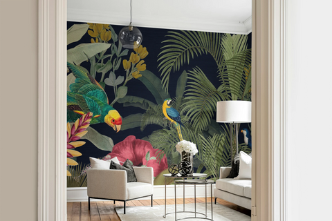 A styled interior room showing a large tropical wallpaper mural with dark navy background, green palm leaves, colorful birds, and pink flowers. The mural is installed behind a light beige armchair, a small round table with flowers, and a white sofa, framed by cream door trim.