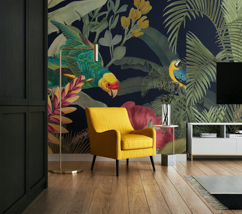 A styled living room featuring a dark navy tropical wallpaper with parrots, palm leaves, and large pink flowers. The room includes a bright yellow armchair, a gold floor lamp, a white side table with a vase, and a black TV mounted on a white wall.