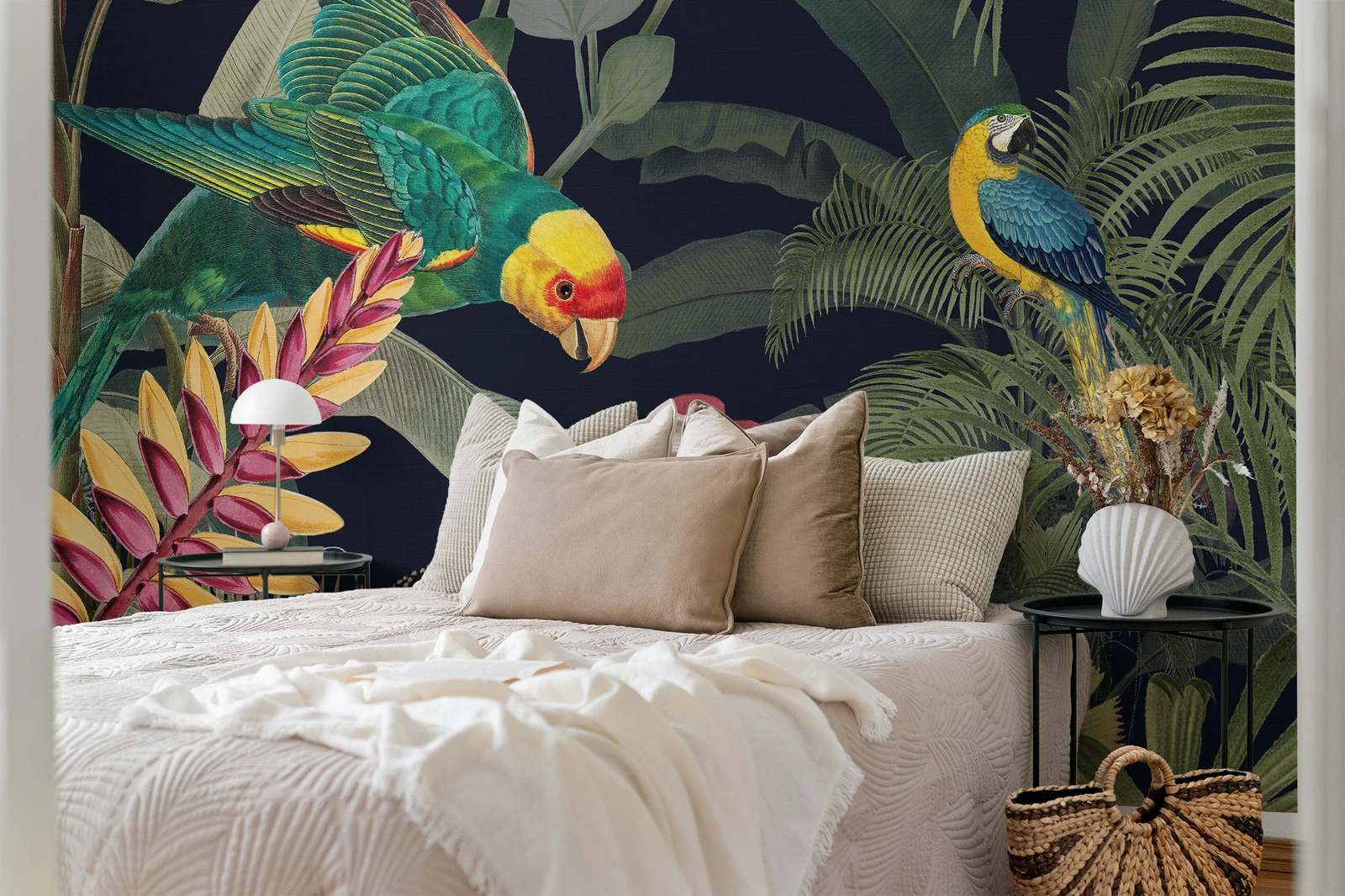 A styled bedroom featuring a bold tropical wallpaper with large green palm leaves and colorful parrots on a dark background. The bed is dressed in cream bedding with beige and textured pillows, flanked by black side tables with a white dome lamp and a white shell vase holding dried flowers.