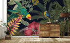 A tropical wallpaper mural featuring a green parrot, a blue-and-yellow macaw, a large pink hibiscus flower, and palm leaves on a dark navy background, shown in a styled room with a wooden dresser, floor lamp, and potted plant.