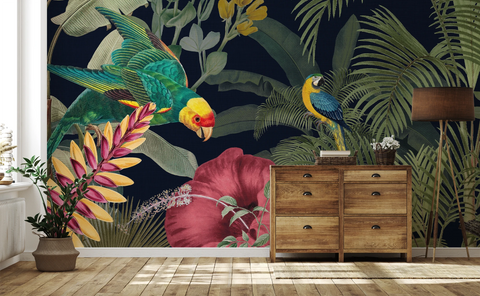 A tropical wallpaper mural featuring a green parrot, a blue-and-yellow macaw, a large pink hibiscus flower, and palm leaves on a dark navy background, shown in a styled room with a wooden dresser, floor lamp, and potted plant.