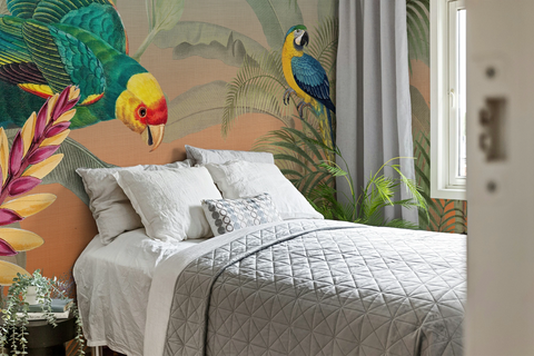 A styled bedroom featuring a large tropical jungle wallpaper mural with colorful parrots and palm leaves. A bed with white linens and a gray quilted comforter is positioned against the wall, next to a window with gray curtains.