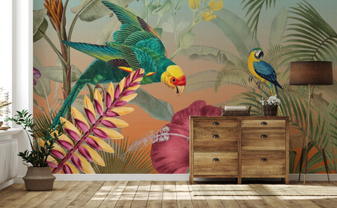 A tropical jungle wallpaper mural featuring a green parrot, a blue-and-yellow macaw, and large palm leaves, shown installed on a wall in a styled room with a wooden dresser, floor lamp, and potted plant.
