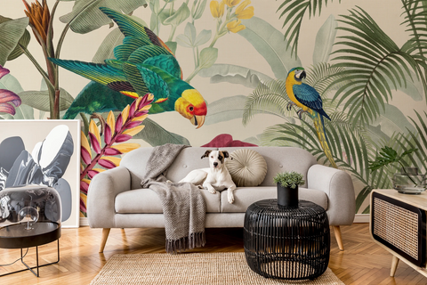 A styled living room featuring a large-scale tropical wallpaper mural with parrots and palm leaves. A light gray sofa sits in the center with a dog lying on it, alongside a black woven side table, a small side table with a mirror, and a vintage-style cabinet.