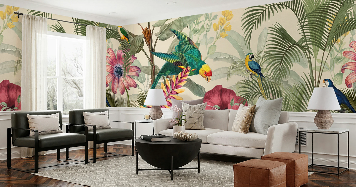 A styled living room featuring a large-scale tropical wallpaper mural with colorful parrots, macaws, and pink flowers on a beige background. The room is furnished with a white sofa, two black armchairs, a black coffee table, side tables with lamps, and brown leather ottomans.