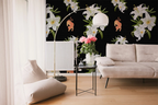 A styled living room featuring a bold black wallpaper with large white lilies and orange tigers, styled with a beige sofa, floor cushions, a gold floor lamp, and a small side table with pink flowers.