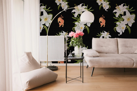 A styled living room featuring a bold black wallpaper with large white lilies and orange tigers, styled with a beige sofa, floor cushions, a gold floor lamp, and a small side table with pink flowers.